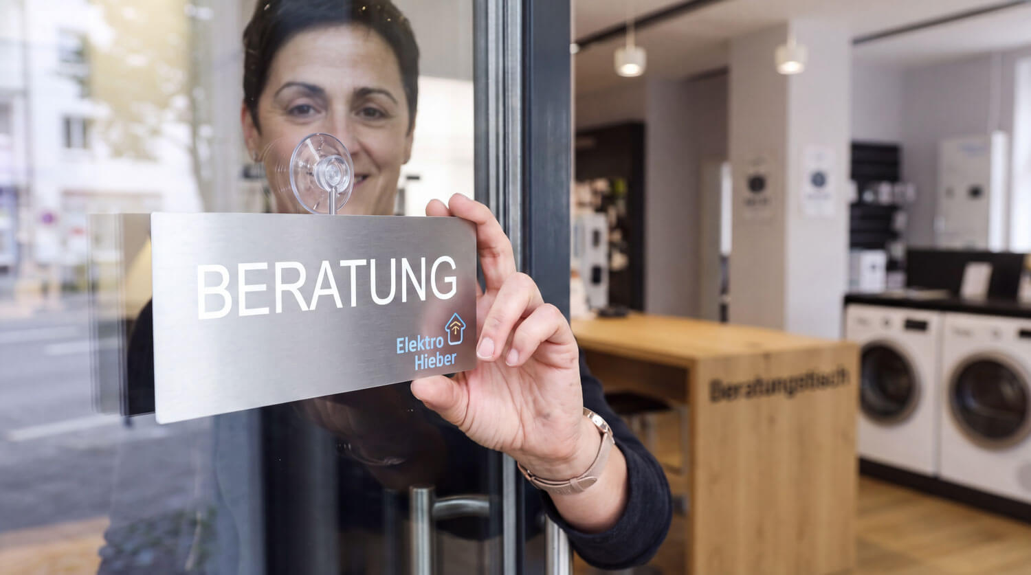 Eine Mitarbeiterin von Elektro Hieber bringt von innen ein Schild mit der Aufschrift Beratung an der Glastür des Ladengeschäfts an.